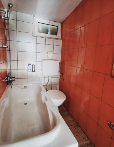 Kupaonica s bijelom kadom i crvenim pločicama na zidovima te malenim prozorom iznad WC školjke.