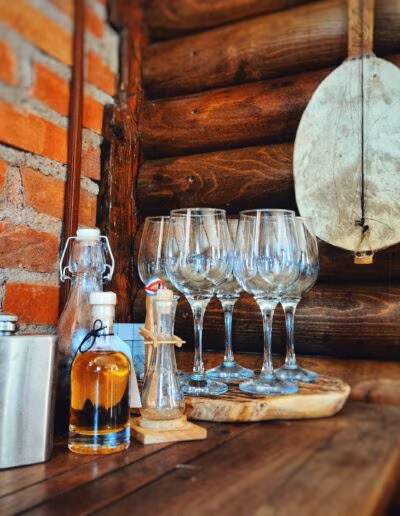 Interijer kuće s drvenim i ciglenim zidovima, na stolu se nalaze staklene boce s pićem, čaše na stalku i metalna pljoska.