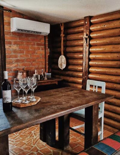 Unutrašnjost rustikalne kuće s drvenim zidovima i stolom na kojem se nalaze boca vina i nekoliko čaša; prozor s crveno-bijelim kariranim zavesama i zidni ukrasi od drva.