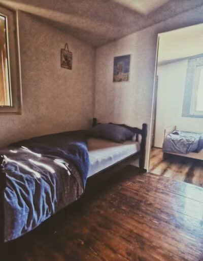 Sobna unutrašnjost s drvenim podom, jednostavnim ležajem i prirodnom rasvjetom kroz prozor, s dodatnom sobom vidljivom u pozadini.