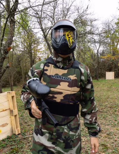 Osoba u maskirnom odijelu s paintball puškom stoji na šumskom terenu, noseći zaštitnu masku s žutim tragovima boje.