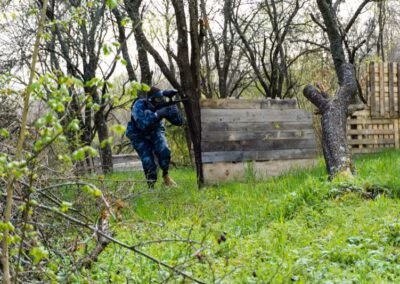 Osoba u maskirnoj odjeći igra paintball skrivena iza drvenog zida u šumovitom području, okružena zelenilom i drvećem.