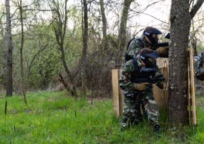 Osobe u kamuflažnim uniformama sudjeluju u igri paintballa u šumovitom području, skrivajući se iza drveća i drvenih barikada.
