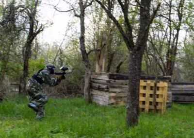 Osoba u maskirnoj uniformi igra paintball na šumskom terenu s drvenim preprekama.