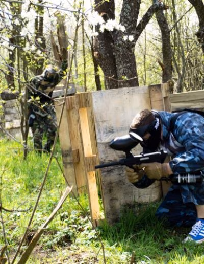 Dvije osobe u maskirnim uniformama sudjeluju u paintball igri skrivajući se iza drvenih prepreka u šumovitom okruženju.