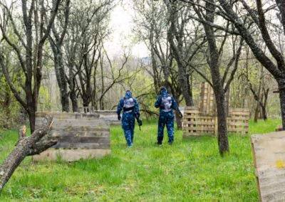 Dvoje ljudi u maskirnim odijelima hodaju kroz šumovito područje s drvenim barikadama, vjerojatno sudjelujući u igri paintballa.