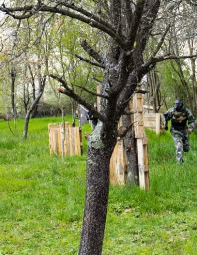 Osoba u maskirnoj odjeći trči kroz šumovito područje s travom i drvenim zaprekama za paintball.
