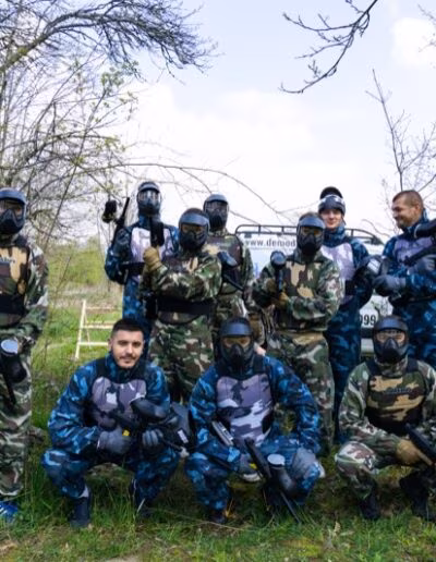 Skupina ljudi u maskirnim odijelima s paintball opremom stoje ispred znaka na kojem piše "www.demod.hr" u šumovitom okruženju.