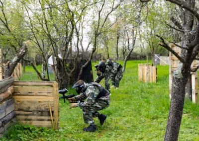 Osobe u maskirnim odijelima igraju paintball na vanjskom terenu s drvenim preprekama usred zelenog okruženja s drvećem.