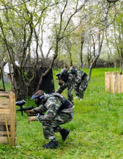Osobe u maskirnim odijelima igraju paintball na vanjskom terenu s drvenim preprekama usred zelenog okruženja s drvećem.