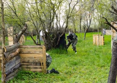 Osobe u maskirnim uniformama sudjeluju u paintball igri na otvorenom prostoru s drvenim barikadama i drvećem.