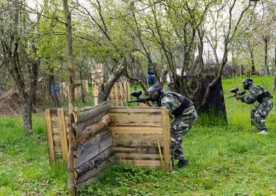 Tri osobe u maskirnim uniformama sudjeluju u igri paintballa u šumovitom području, skrivajući se iza drvenih prepreka.