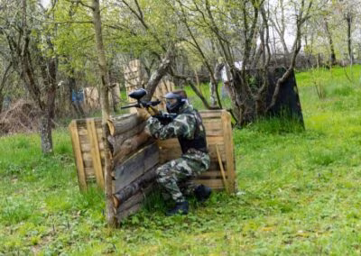 Osoba u maskirnoj uniformi i zaštitnim naočalama igra paintball, čučeći iza barikade u šumovitom području.