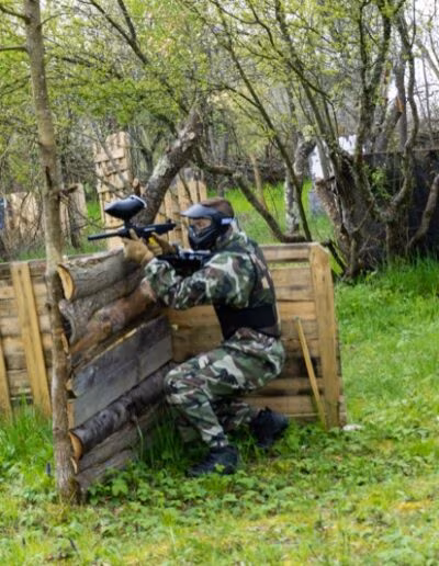 Osoba u maskirnoj uniformi i zaštitnim naočalama igra paintball, čučeći iza barikade u šumovitom području.