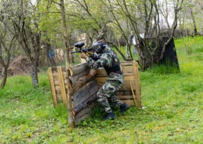 Osoba u maskirnoj odjeći igra paintball, skriva se iza drvenog zaklona u šumovitom području, s puškom i zaštitnom maskom.