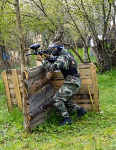 Osoba u maskirnoj odjeći igra paintball, skriva se iza drvenog zaklona u šumovitom području, s puškom i zaštitnom maskom.