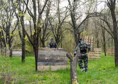Osobe u maskirnim odorama igraju paintball u šumovitom okruženju, skrivajući se iza drvenih prepreka.