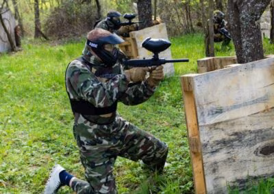 Osoba u maskirnoj odjeći igra paintball, čučeći iza drvene barikade u šumi, s paintball puškom u ruci.