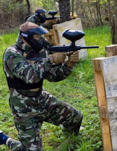 Osoba u maskirnoj odjeći igra paintball, čučeći iza drvene barikade u šumi, s paintball puškom u ruci.