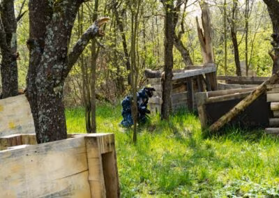 Osoba u maskirnoj odjeći skriva se iza drvene barikade u šumskom okruženju s drvenim preprekama i gustim zelenilom.