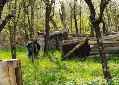Osoba u maskirnoj odjeći i zaštitnoj maski skriva se iza drvene ograde u šumi okruženoj drvećem i zelenom travom.