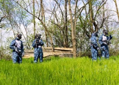 Četiri osobe u opremi za paintball stoje na zelenoj livadi ispred šumovitog područja.