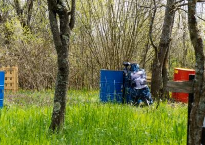 Osoba u šarenom maskirnom odijelu skriva se iza plavog metalnog bureta u šumovitom području, vjerojatno sudjelujući u paintball igri.