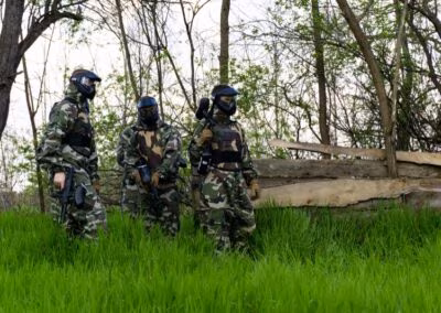 Tri osobe u maskirnim uniformama i zaštitnim maskama igraju paintball u šumi s visokim travama i drvenim preprekama.