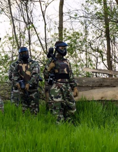 Tri osobe u maskirnim uniformama i zaštitnim maskama igraju paintball u šumi s visokim travama i drvenim preprekama.