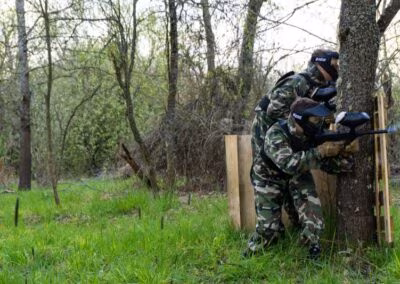 Dvije osobe u maskirnim uniformama igraju paintball u šumovitom području, skrivajući se iza stabla i drvenog zaklona.