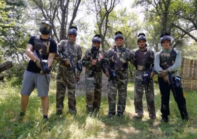 Šest osoba u kamuflažnim odijelima i zaštitnim maskama drže paintball puške, stojeći na travnatom području okruženom drvećem.