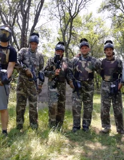 Šest osoba u kamuflažnim odijelima i zaštitnim maskama drže paintball puške, stojeći na travnatom području okruženom drvećem.
