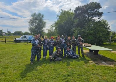 Grupa ljudi u maskirnim odijelima i s paintball opremom stoji na travnatom terenu, uz stol i drveće u pozadini.