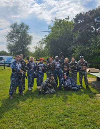 Grupa ljudi u maskirnim odijelima i s paintball opremom stoji na travnatom terenu, uz stol i drveće u pozadini.