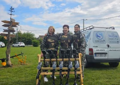 Tri osobe u maskirnim uniformama stoje ispred police s paintball puškama i kombija, na otvorenom prostoru s travom i drvenim putokazom u blizini.