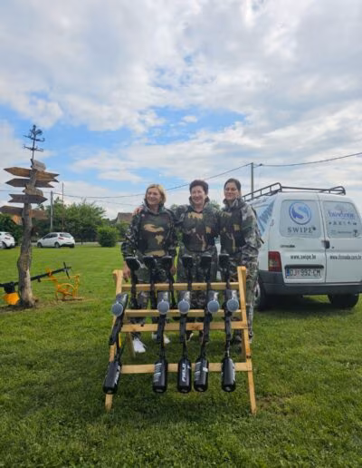 Tri osobe u maskirnim uniformama stoje ispred police s paintball puškama i kombija, na otvorenom prostoru s travom i drvenim putokazom u blizini.