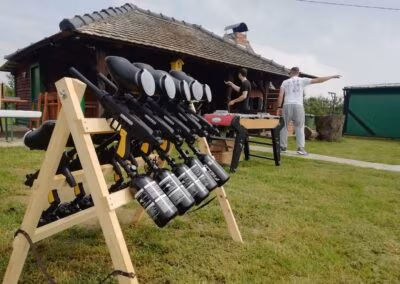 Kuća od drva s krovom od crijepa stoji u pozadini na travnatoj površini, dok su u prvom planu postavljeni paintball pištolji na drvenom postolju; dvije osobe stoje u blizini stola za stolni nogomet.