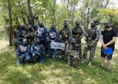 Grupa ljudi u paintball opremi stoji na travnatom području okruženom drvećem; neki nose zelene, a neki plave kamuflažne uniforme, s natpisom "Demode" ispred njih.