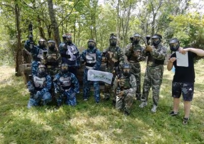 Grupa ljudi u paintball odorama stoji na travnatoj površini ispred drvene strukture, okružena drvećem, neki držeći paintball puške.