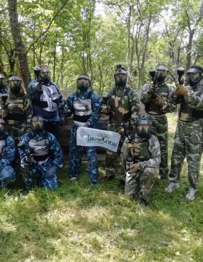 Grupa ljudi u maskama i odijelima za paintball pozira na livadi okruženoj drvećem, držeći paintball puške, ispred znaka s natpisom 'Demode.'