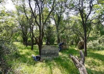 Ljudi igraju paintball u šumovitom području s drvenim preprekama i kotačima, okruženi gustom vegetacijom i drvećem.