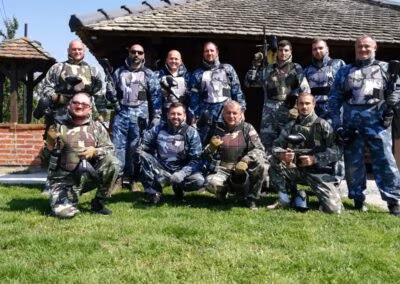 Grupa muškaraca u maskirnim odijelima za paintball, pozirajući ispred rustikalne drvene kućice na travnjaku.