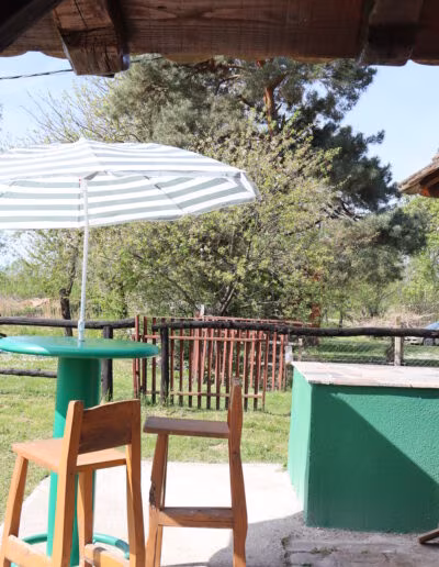Terasa s drvenim stolcima i zelenim stolom pod bijelim prugastim suncobranom, okružena drvećem i ogradom na seoskoj lokaciji.