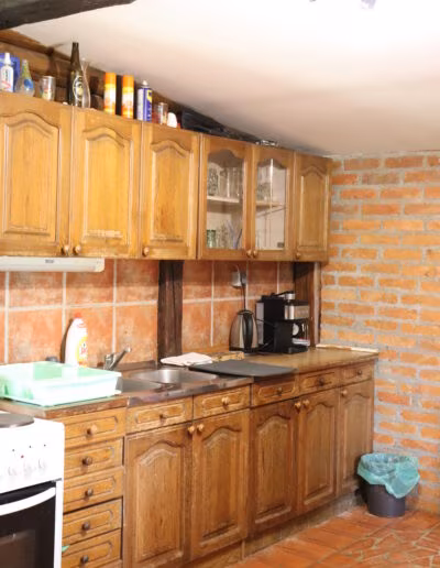 Kuhinja s drvenim ormarićima, otvorenim policama s čašama, smeđim pločicama na zidovima i podu, i kućanskim aparatima poput kuhala za vodu i aparata za kavu na radnoj ploči.