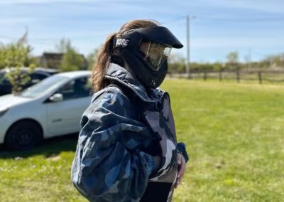 Osoba u kamuflažnom prsluku s maskom za zaštitu lica stoji na travnatom području pored automobila, pod vedrim nebom.