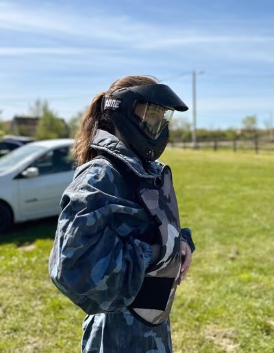 Osoba u kamuflažnom prsluku s maskom za zaštitu lica stoji na travnatom području pored automobila, pod vedrim nebom.