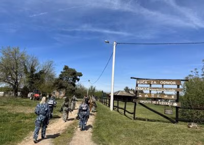 Grupa ljudi u maskirnim uniformama hoda poljskom stazom pored drvenog znaka na kojem piše 'Kuća za odmor Demode', s vedrim nebom i zelenilom u pozadini.