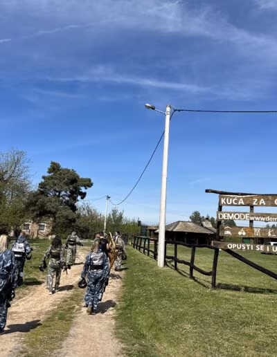 Grupa ljudi u maskirnim uniformama hoda poljskom stazom pored drvenog znaka na kojem piše 'Kuća za odmor Demode', s vedrim nebom i zelenilom u pozadini.