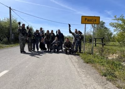 Grupa ljudi u maskirnim odorama s paintball puškama stoji na cesti pored znaka naselja Paučje, okružena prirodom i plavim nebom.