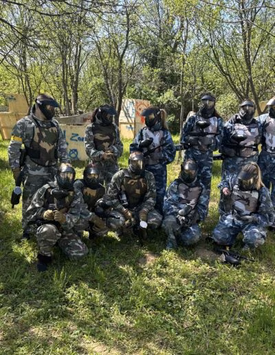 Grupa ljudi u maskirnim uniformama pozira na paintball terenu okruženom drvećem i drvenim preprekama.
